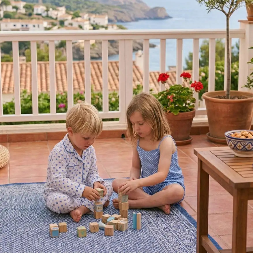 Deux enfants en pyjama et une robe bleue jouent avec des blocs en bois sur un tapis à motifs.